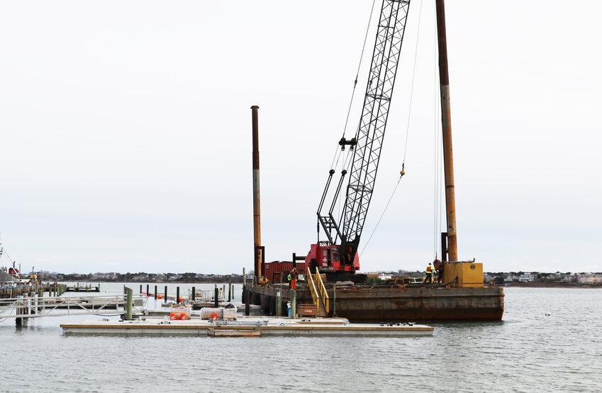 A crane on a barge is replacing the floating wooden docks at the town pier with concrete ones.