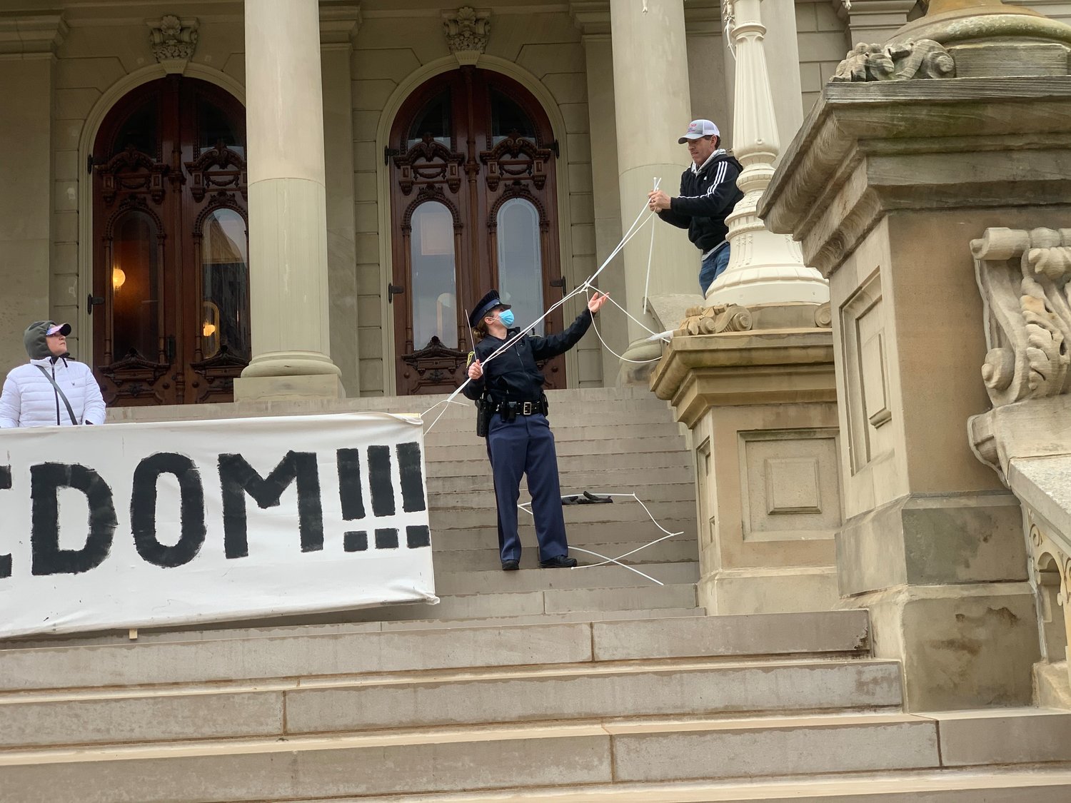 At least one Michigan State Police officer helped demonstrators hang a “Freeedom!!!” sign over the steps of the State Capitol building today.