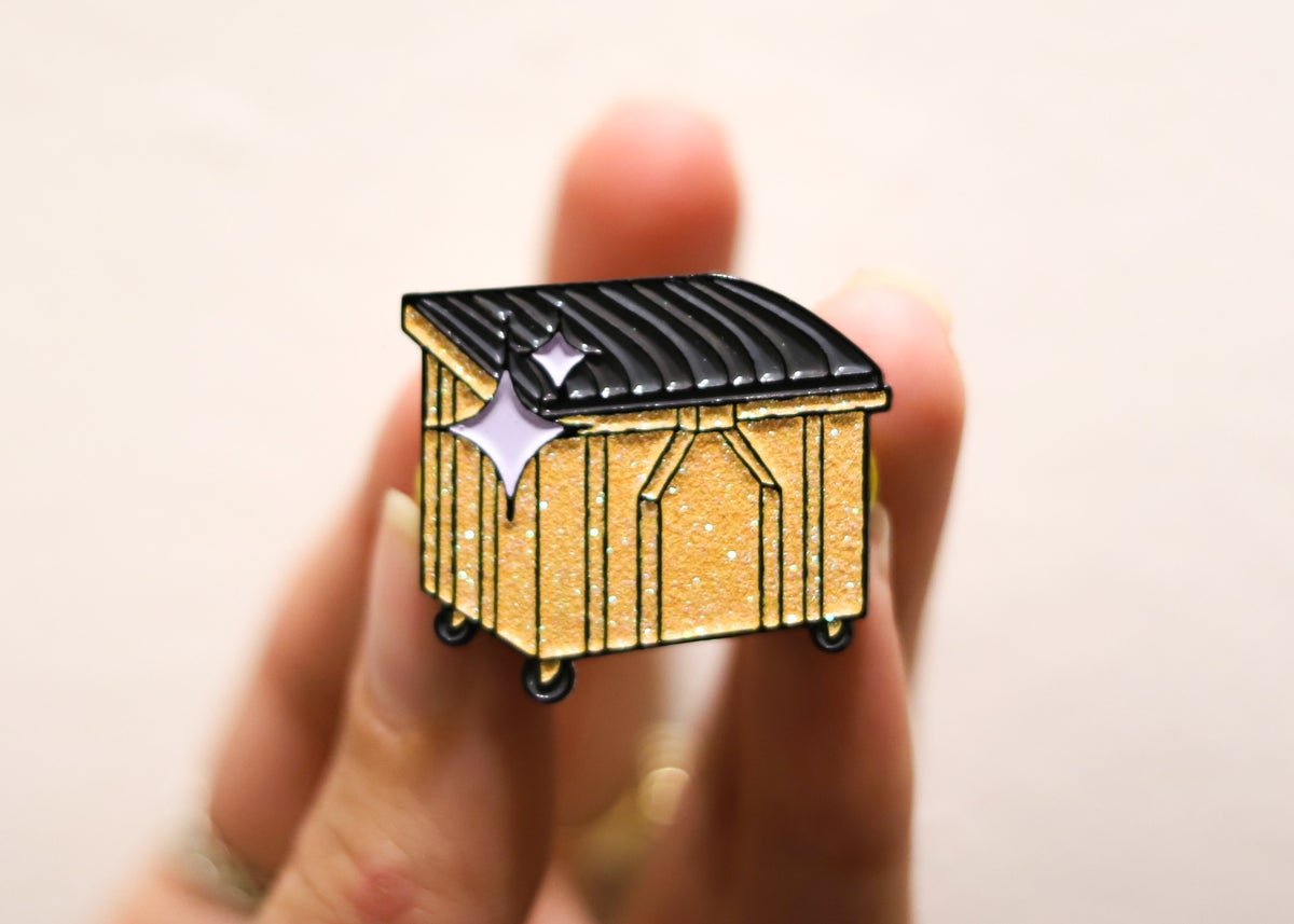 An enamel pin based on Mac's Bar's infamous golden dumpster.