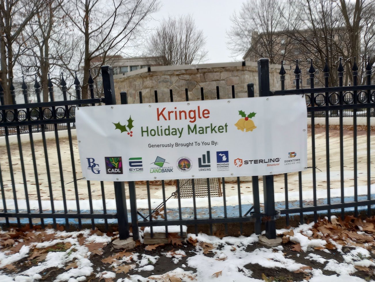 A banner hung on the fence around the fountain in Reutter Park in downtown Lansing announcing the Kringle Holiday Market. The market features eight wooden structures and various vendors now through Dec. 17.