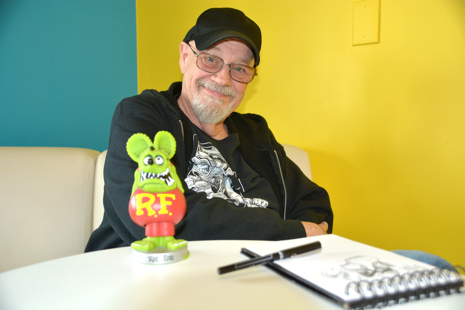 Local artist Dennis Preston poses with his trusty sketch pad and a figurine of Ed “Big Daddy” Roth’s Rat Fink caricature, a major inspiration for his own art.