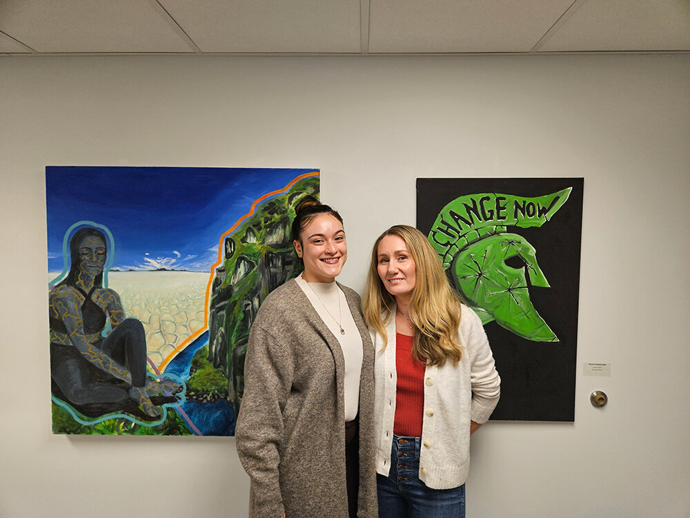 Mya Trevino (left) and Claire Redmer of Small Talk Children’s Advocacy Center stand with art by Rachel Nanzer and Ryan Holmes, on display at the Lansing Art Gallery through Oct. 28.