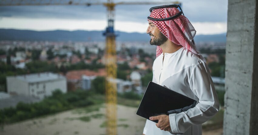 An Arab man wearing a kaffiyeh holds a black book and looks towards the skyline of Dubai.