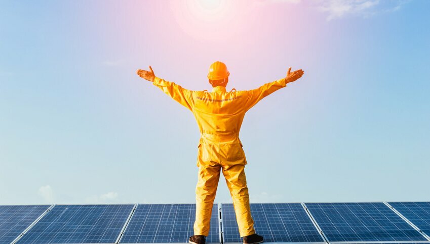 Man in yellow work gear stands arms wide on solar panels.