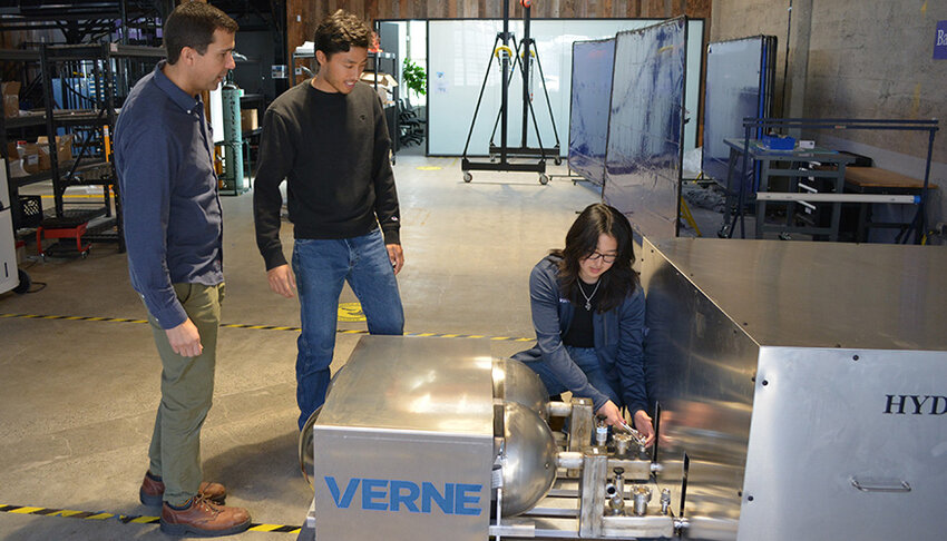 The high-pressure cryogenic heat exchanger-based system used for this work is shown to the right of the picture. From left to right, Nick Killingsworth, a mechanical engineer at LLNL, and Telis Athanasopoulos Yogo, a Verne mechanical engineer, watch as Kara Zhang, a Verne process engineer, installs a vacuum pressure gauge on the system.
