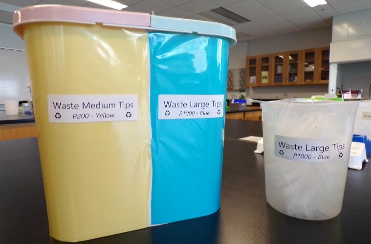 This image, taken inside a Georgia Tech laboratory, displays a set of clearly labeled recycling bins used for pipette tip disposal. The bins are part of the university’s Tip-Cycle program, an innovative lab sustainability initiative aimed at reducing plastic waste. The containers are color-coded and labeled by pipette tip size: a yellow bin for "Waste Medium Tips (P200 – Yellow)," a blue bin for "Waste Large Tips (P1000 – Blue)," and an additional translucent container also designated for large pipette tips. The background reveals a modern lab environment, reinforcing Georgia Tech’s commitment to integrating sustainability into cutting-edge research spaces.