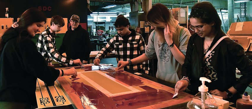 High schoolers, from left, Victoria Wall, Weston Ballantine, Avery Miller, Brayden Metz and Alex Osterkamp get hands on during their tour of Signcraft learning how to make a screen from employee Renee Reddington. Not pictured, Derek Engle.