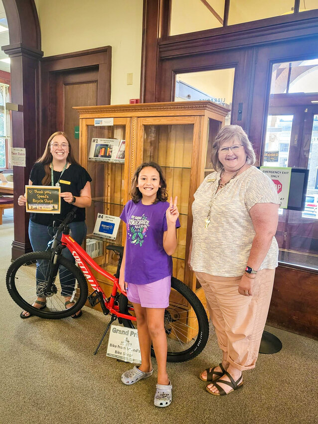 Librarian Cheyenne Puetz (Left), Super Reader Nadia Sletten (Center), and Library Director Michelle Tryggestad (Right) pause for a commemorative photo, as they celebrate Nadia&rsquo;s grand prize win for the Summer Learning Challenge. Her amazing prize earned was a brand-new bike from Speed&rsquo;s Bike Shop! Way to go, Nadia!