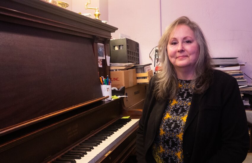 Dana Tock sitting beside her 100 year old upright grand piano that her students have been learning on since 1989.