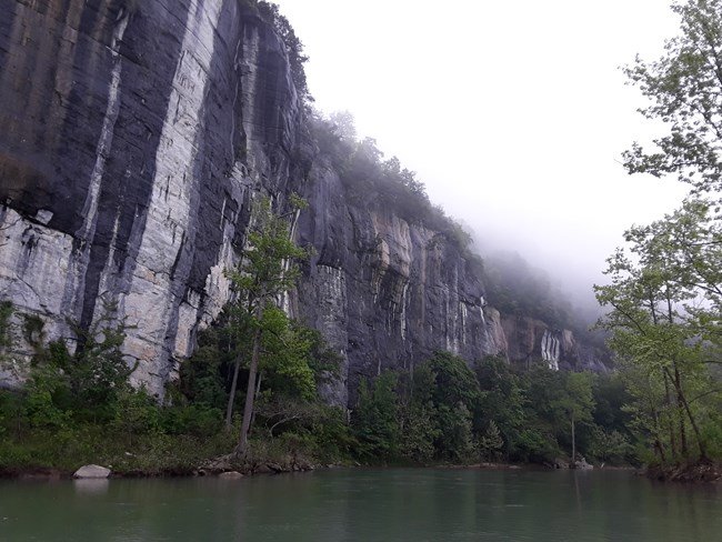 A limestone bluff juts out into the Buffalo River. The bluff has several solution cavities at its base, where the limestone meets the river. The rock face has colors of white, gray, and light orange.