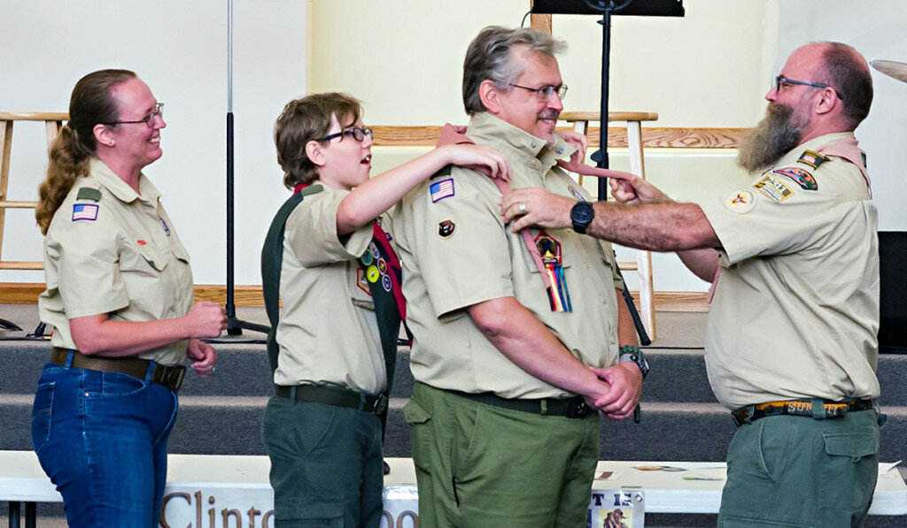Photo Ron Armstrong — 
Woodbadge Course Director for C3-138-19 James Howard along with Ed Cicenas’s son Zander place the neckerchief bearing the Woodbadge Tartan around his neck while his wife Krystal looks on. Cicenas received his Woodbadge beads and neckerchief at a special ceremony at Lane Christian Church on Monday. His son also achieved a special honor that night, receiving his First Class rank as a member of Clinton’s BSA Troop 1142.