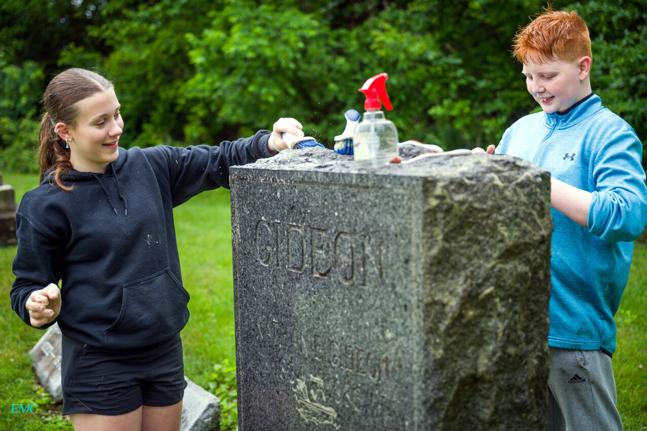 This year, 120 eighth-grade social studies students from CJHS, under the guidance teacher Kelby McMath, continue “The Good Cemeterian Project,”  Here, Aaliyah Osborne (left), and Max Marlow (right), work to clean the gravestone of a member of the Gideon family, a name out of the history of Clinton.
