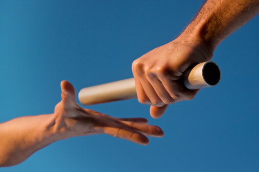 Low angle close-up of two hands passing a relay baton, with focus on the hand holding the baton, against a clear blue sky. Warm tones highlight the fact that the image was shot at sunset. Horizontal format.
