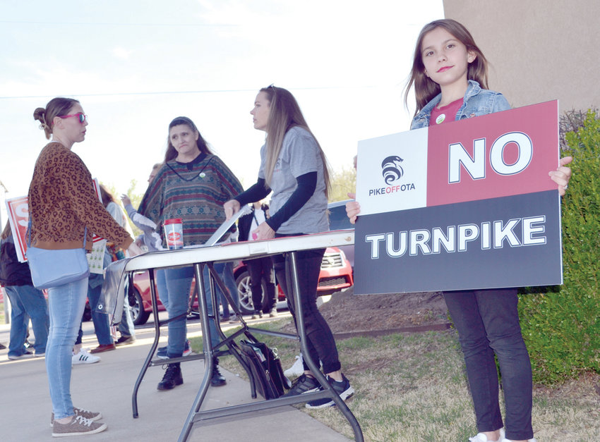 Ashlynn Duffey of Norman stands in front of Newcastle Public Schools&rsquo; administration building Monday evening, and in front of her mother, Mindy, and her sister (next to Mom), Jaidyn, with a Pike Off OTA sign, just before a meeting of the Turnpike Authority with Newcastle residents.