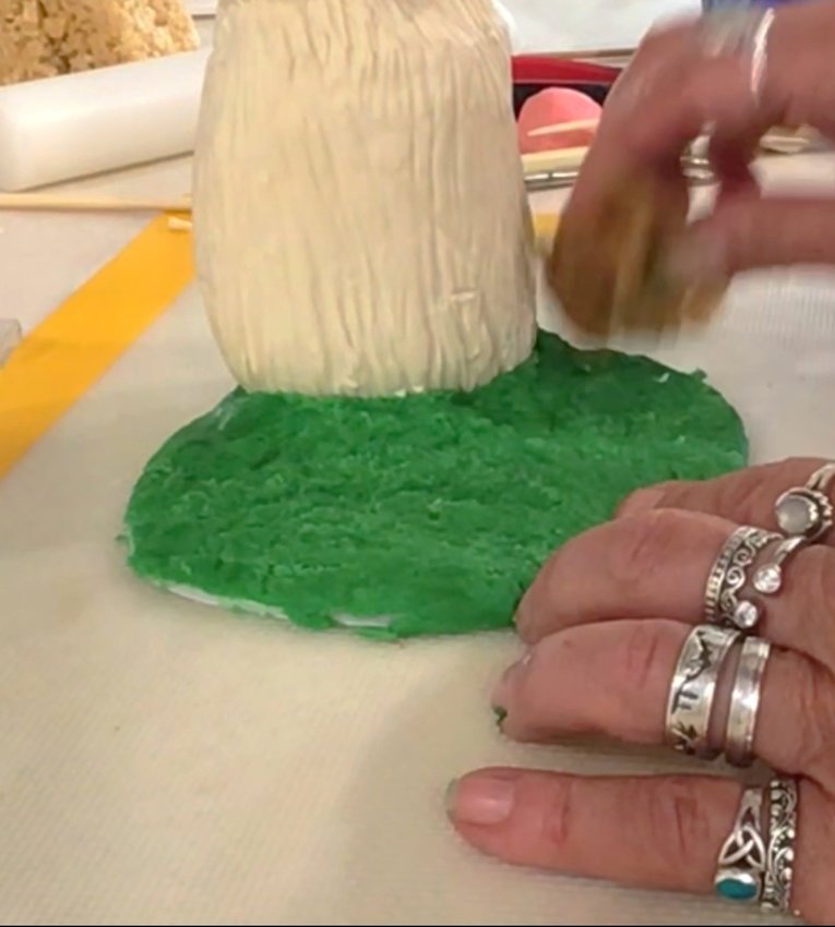Attach head to body using wooden dowel. Smooth in fondant on neck to solidify. Attach body to FoamCore board with piping gel. Cover the rest of the board in green fondant to look like grass.