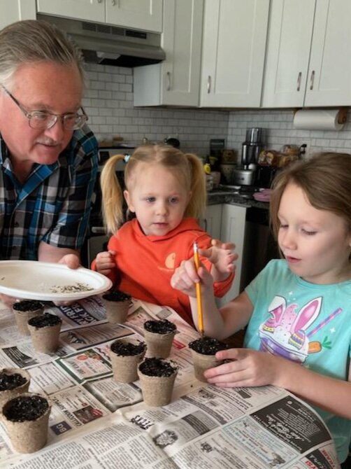 Me with granddaughters Gwendolyn (center) and Alayna (right) planting marigold seeds in my kitchen. Using a pencil to make holes in the soil to let the seeds in.