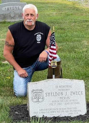 Keith Herbert kneels besides the Sheldon J. Zweck memorial at the Liberty Cemetery as part of the memorial motorcycle poker run August 14th.
