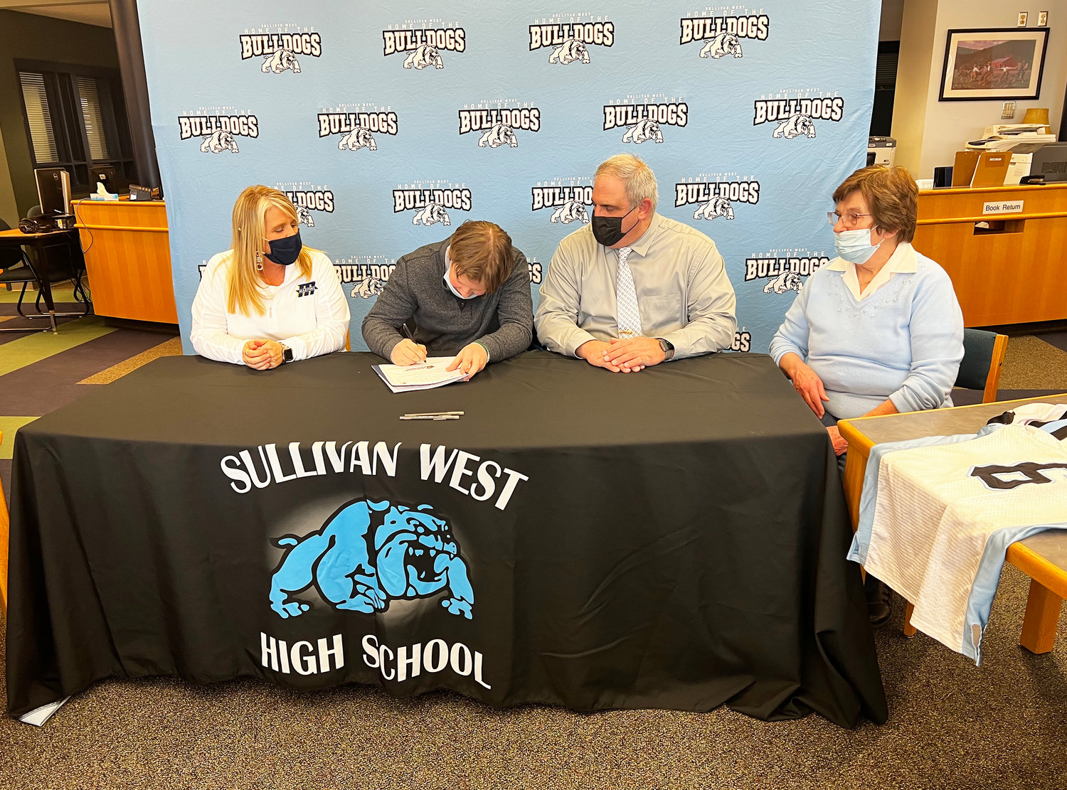 Sullivan West’s Gavin Hauschild pens the Division III Celebratory signing form making it official that he will be attending Massachusetts Maritime Academy to play football and pursue his intended path towards a career in Emergency Management. He is flanked by his mother Dawn, his father John and grandmother Barbara.