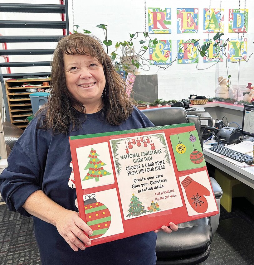 Wednesday afternoon, Dec. 6, Joni Spalding, the new children's librarian at Boonslick Regional Library, holds a poster for a Saturday, Dec. 9 event featuring National Christmas Card Day. Spalding, originally from Sedalia and a retired educator, took the position in October.   Photo by Faith Bemiss-McKinney | Democrat