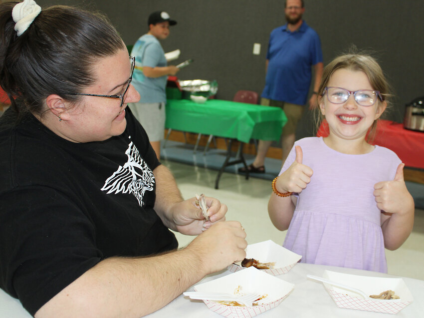 Two thumbs up – Lyla Miller gives the chicken wings two thumbs up during the Saturday, July 20 barbecue cook-off for Operation Christmas Child at the Eugene Christian Church. With her is her mom Samantha.