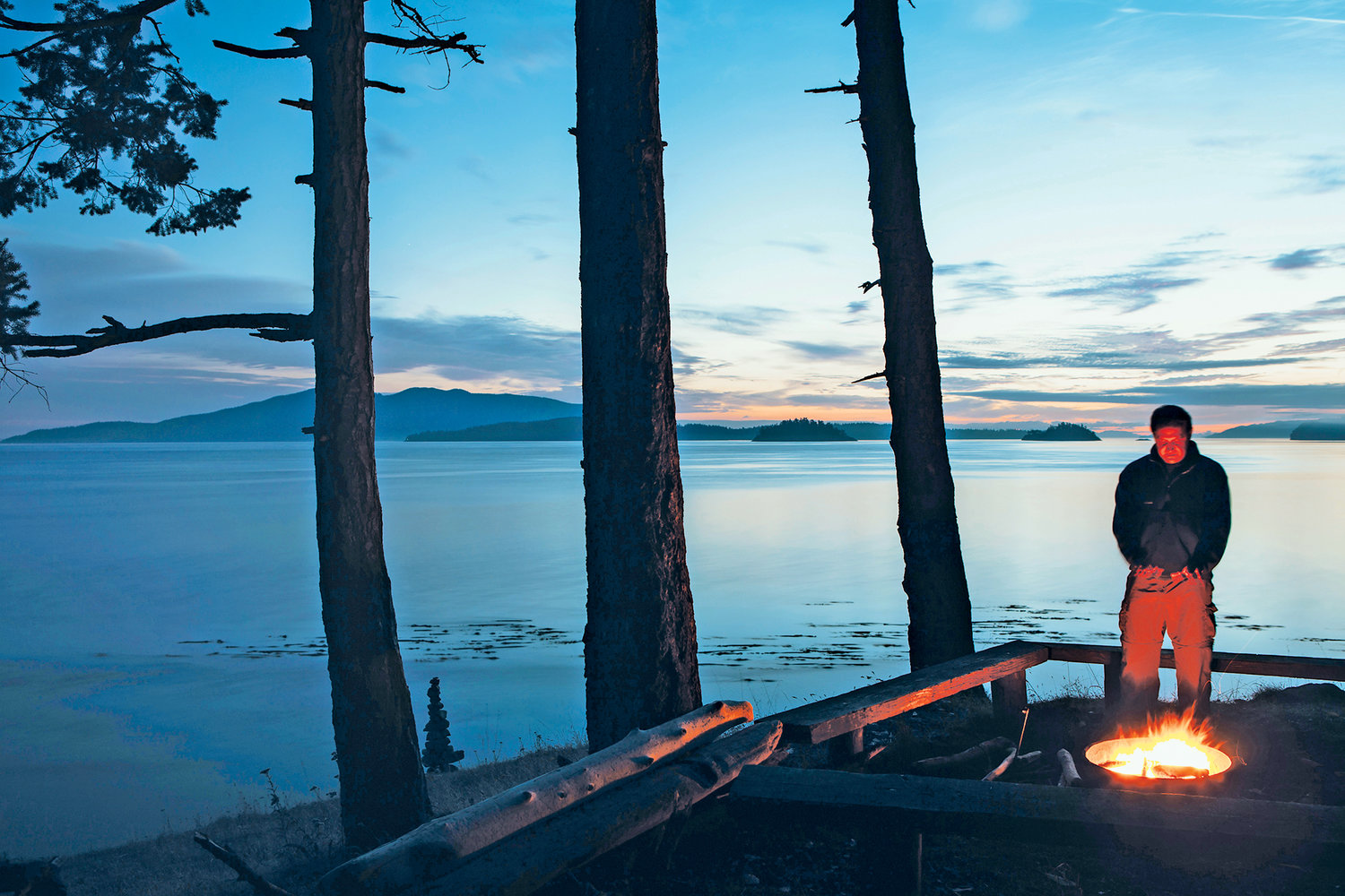 Man standing by campfire at dusk, San Juan Islands in the distance, Washington, USA.